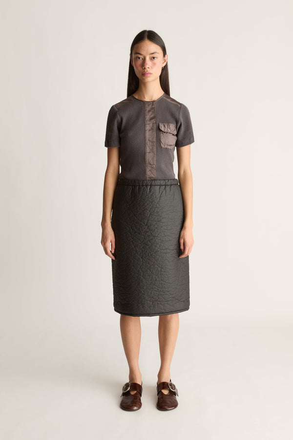 A model stands against a light gray studio background wearing a dark gray short-sleeve top with a textured vertical chest panel and matching dark gray skirt, paired with brown loafers.