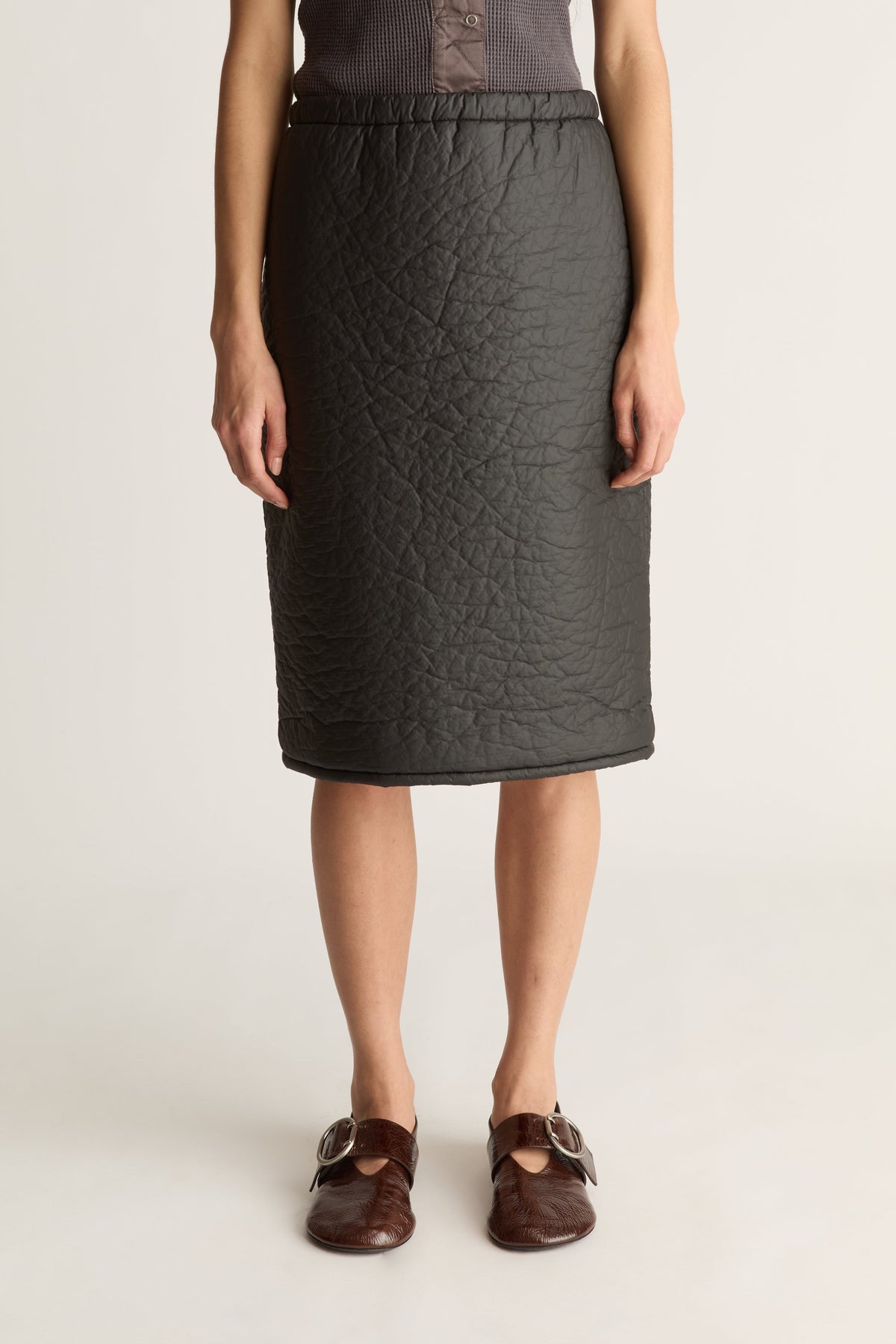 A model wearing a dark gray quilted pencil skirt with an elastic waistband, paired with brown leather mule sandals, against a plain light gray studio background.