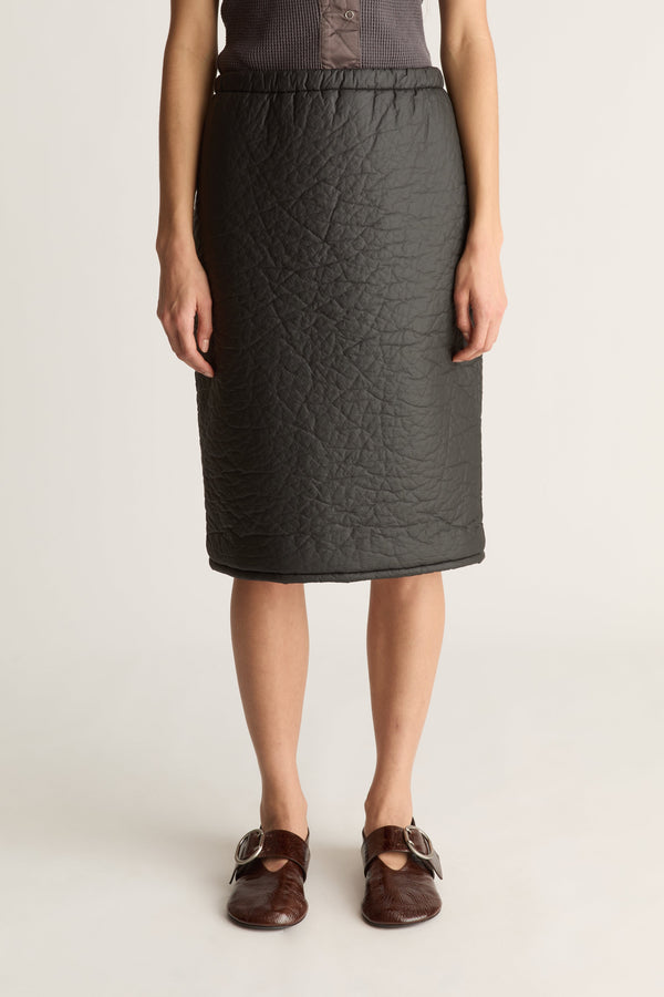 A model wearing a dark gray quilted pencil skirt with an elastic waistband, paired with brown leather mule sandals, against a plain light gray studio background.