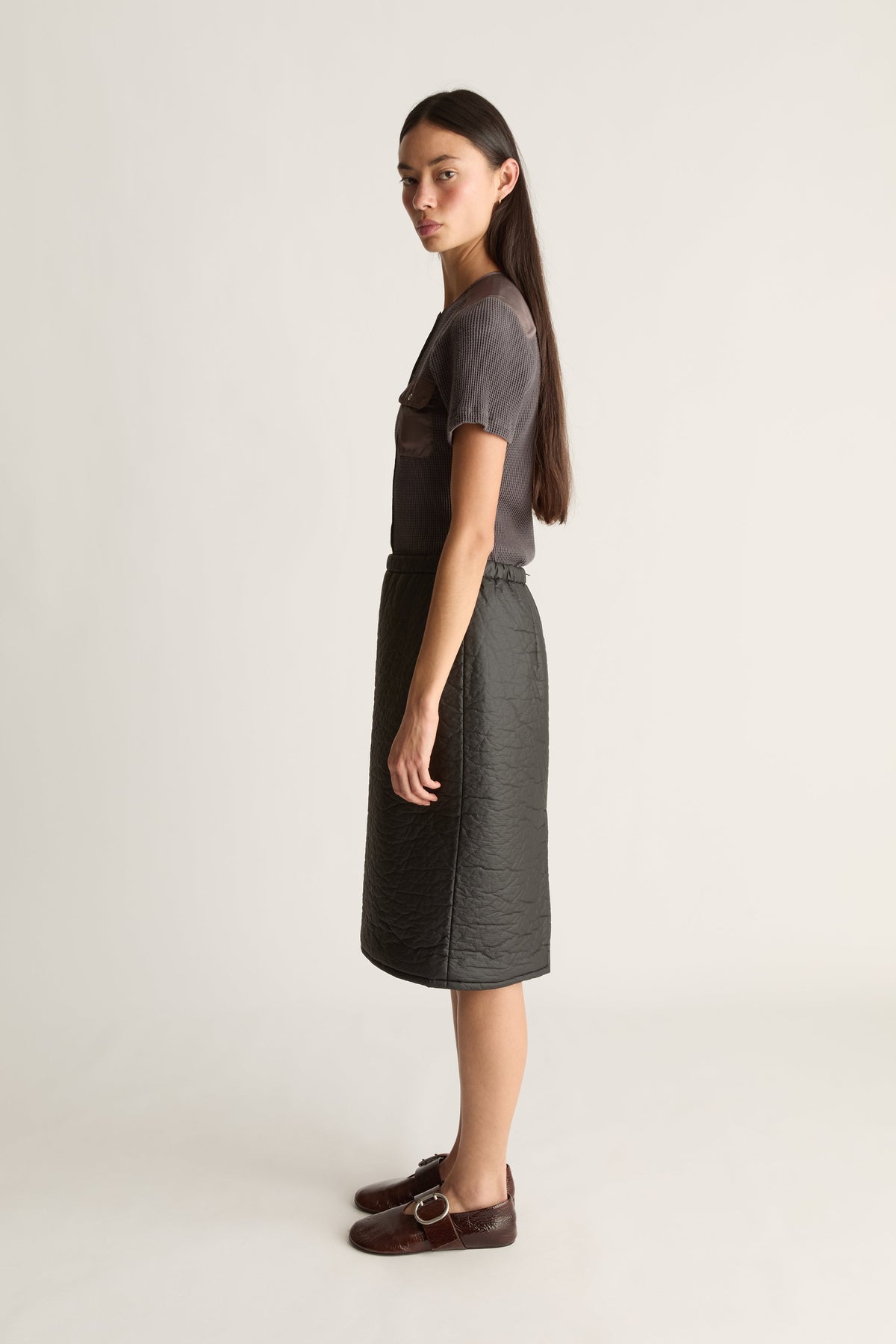 A woman stands in profile wearing a dark gray short-sleeve top and a dark gray midi skirt, paired with dark brown loafers, against a plain light gray studio background.