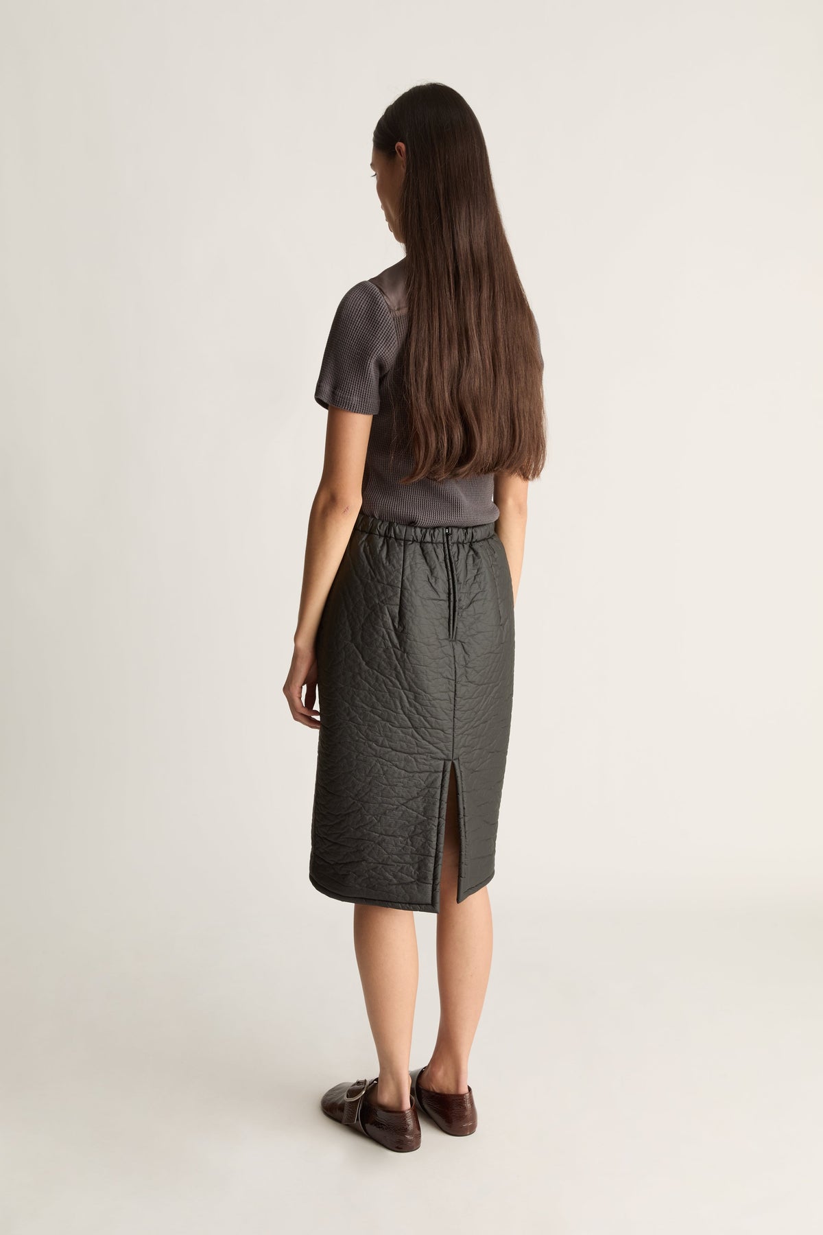 Back view of a woman wearing a  dark gray short-sleeve top and a dark gray textured midi skirt  paired with dark brown loafers, against a plain light gray studio background.