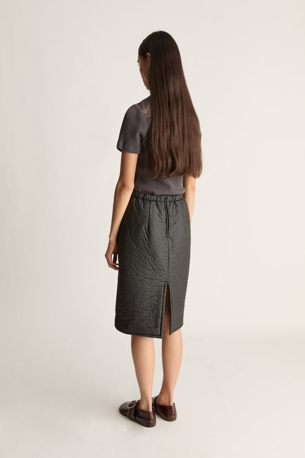 Back view of a woman wearing a  dark gray short-sleeve top and a dark gray textured midi skirt  paired with dark brown loafers, against a plain light gray studio background.