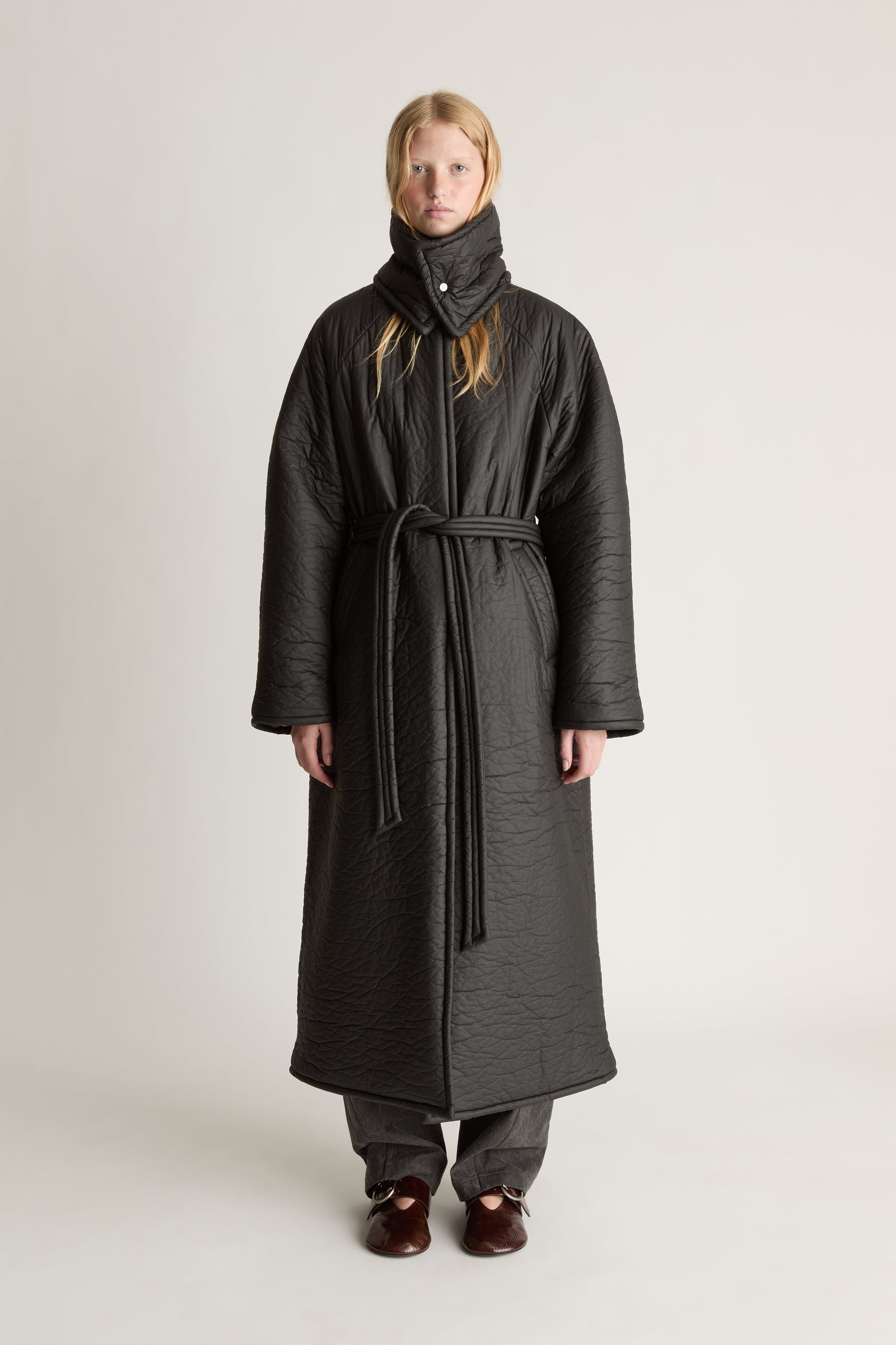 Person facing the camera wearing a long black puff fabric  coat on a plain background