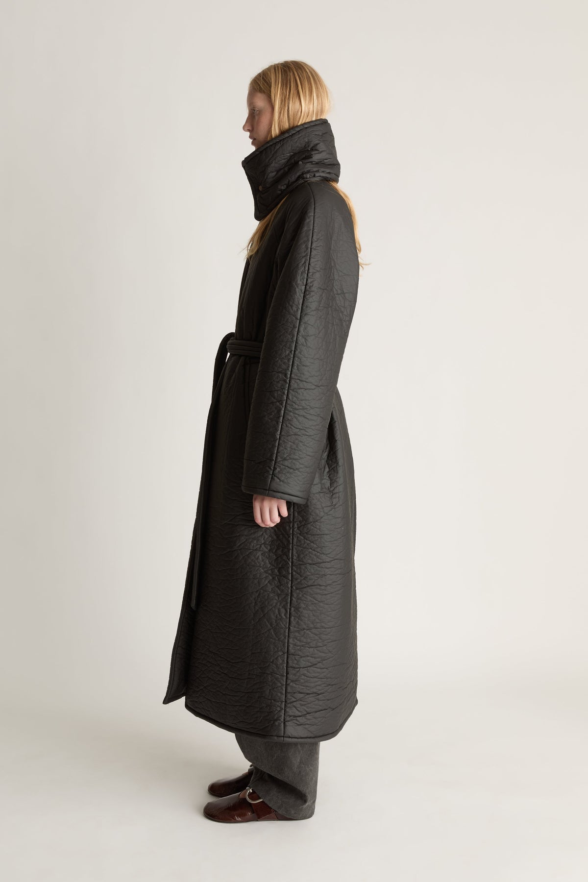 Side view image of a woman wearing a full lenght black parker coat with a waist tie and against studio background
