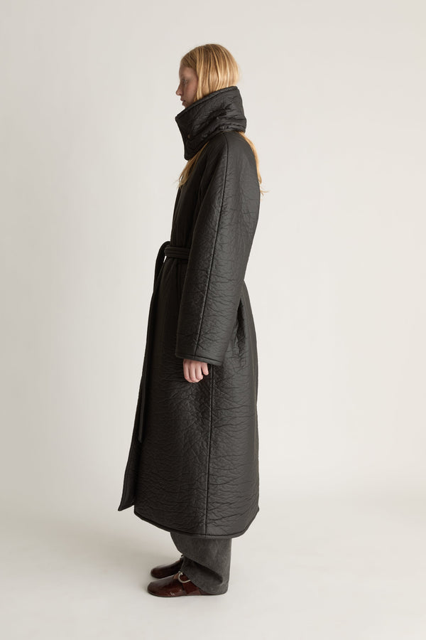 Side view image of a woman wearing a full lenght black parker coat with a waist tie and against studio background