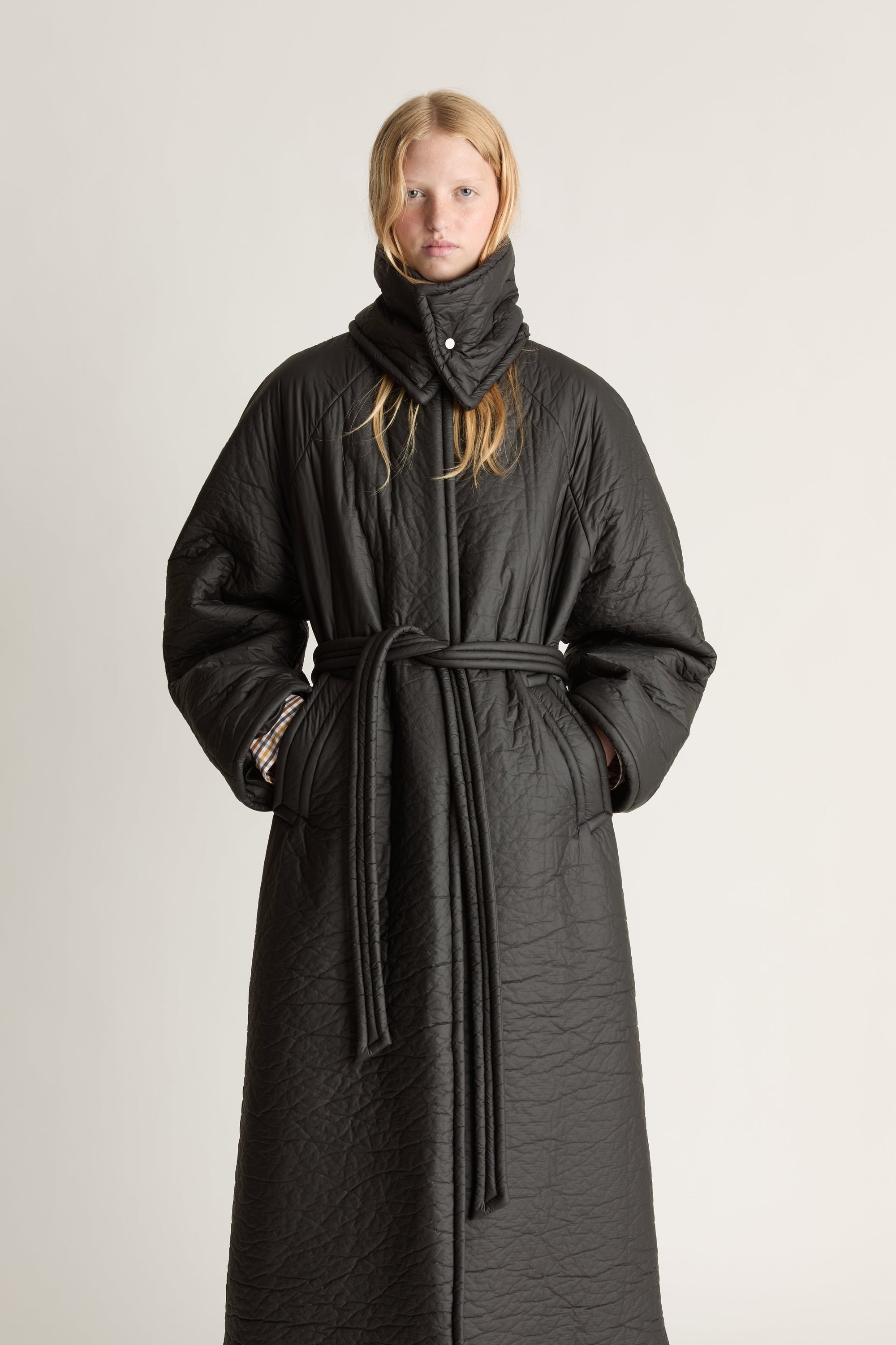 Close up of a person wearing a long black coat with a funel neck and waist tie on a plain background