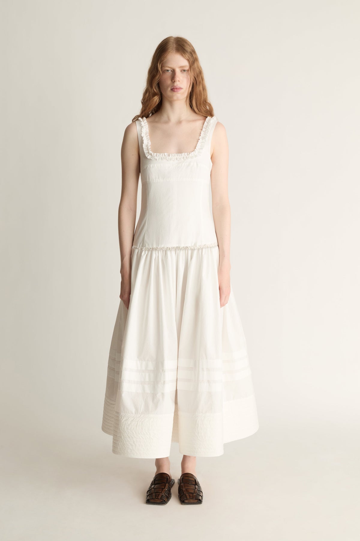 A woman stands against a plain light beige studio background wearing a sleeveless cream dress with a square neckline, delicate lace trim at the shoulders and waist, and a tiered skirt. She is wearing dark brown loafers.