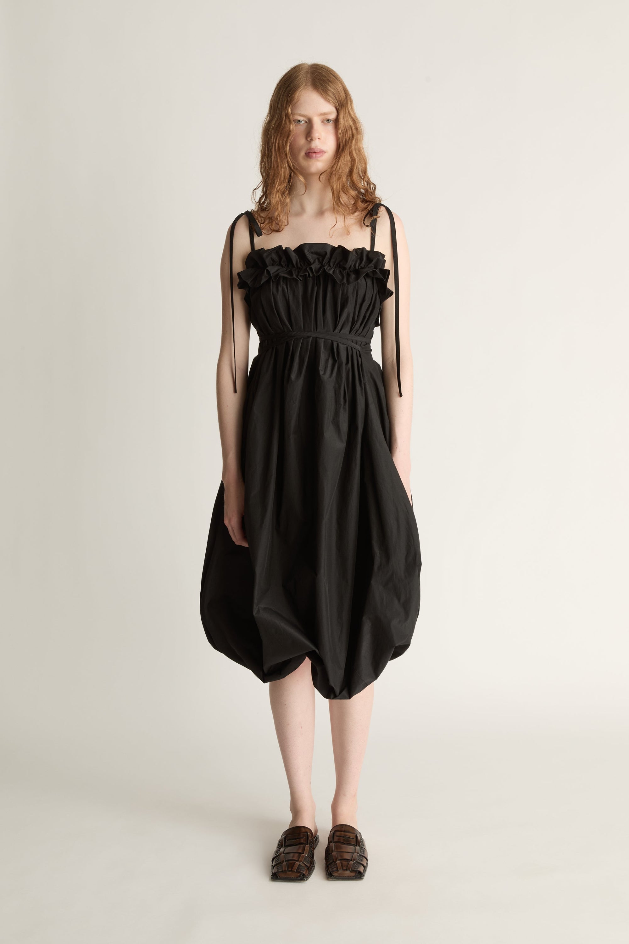 A woman facing the camera wearing a black bubble style dress with black tie straps and brown shoes  against a white studio background.