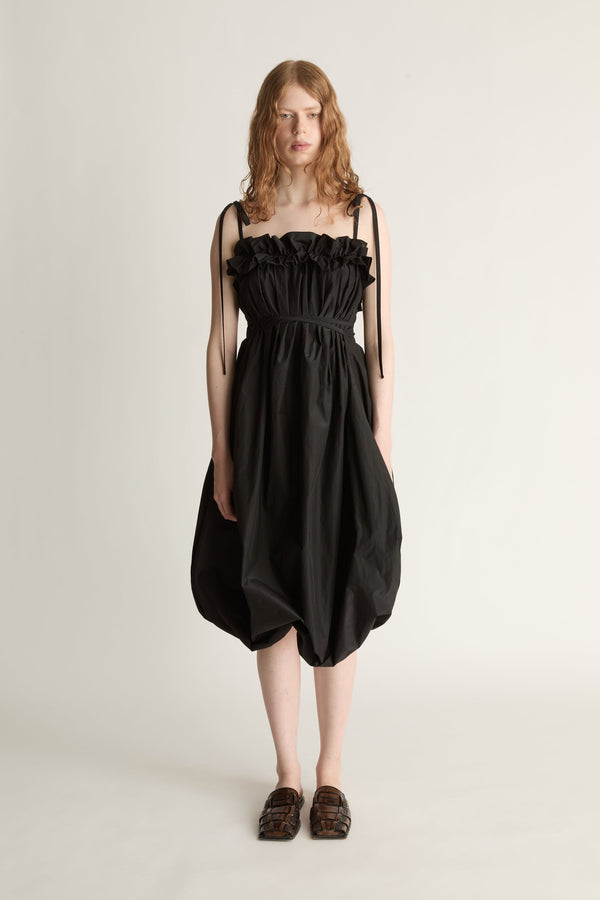 A woman facing the camera wearing a black bubble style dress with black tie straps and brown shoes  against a white studio background.