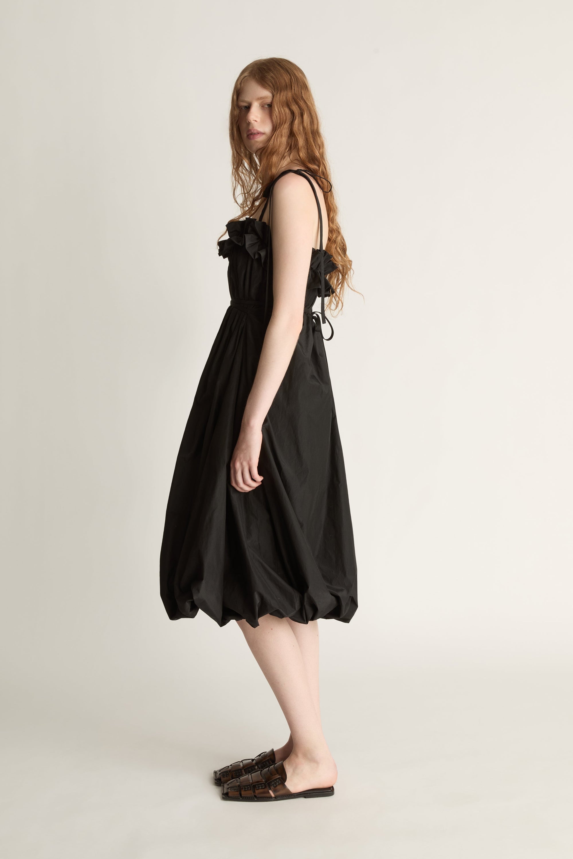 Side view of a woman wearing a black midi length dresss and brown shoes against a white studio background
