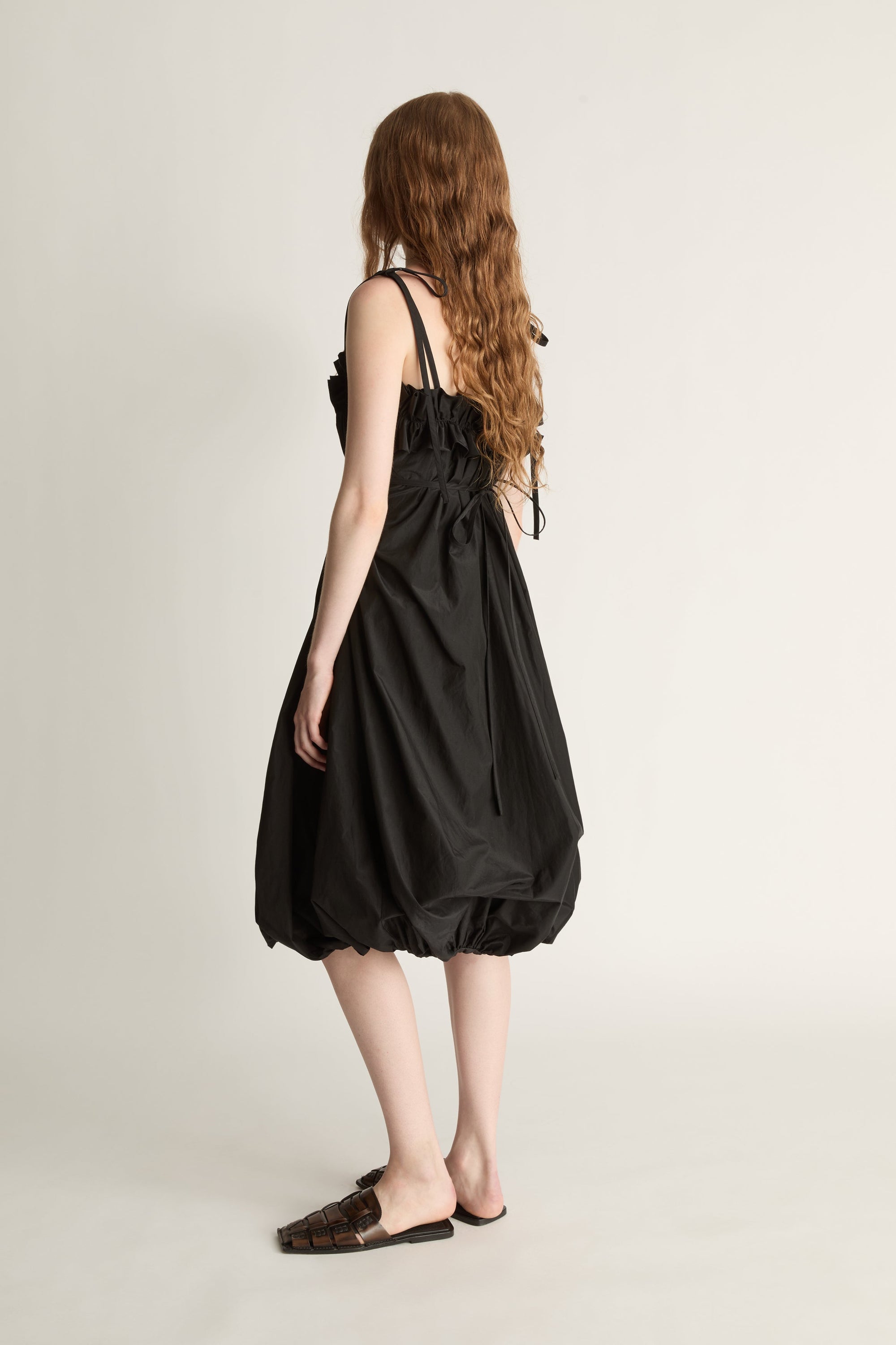 Back view image of a woman wearing a black bubble-style midi dress and brown shoes against a white studio background.