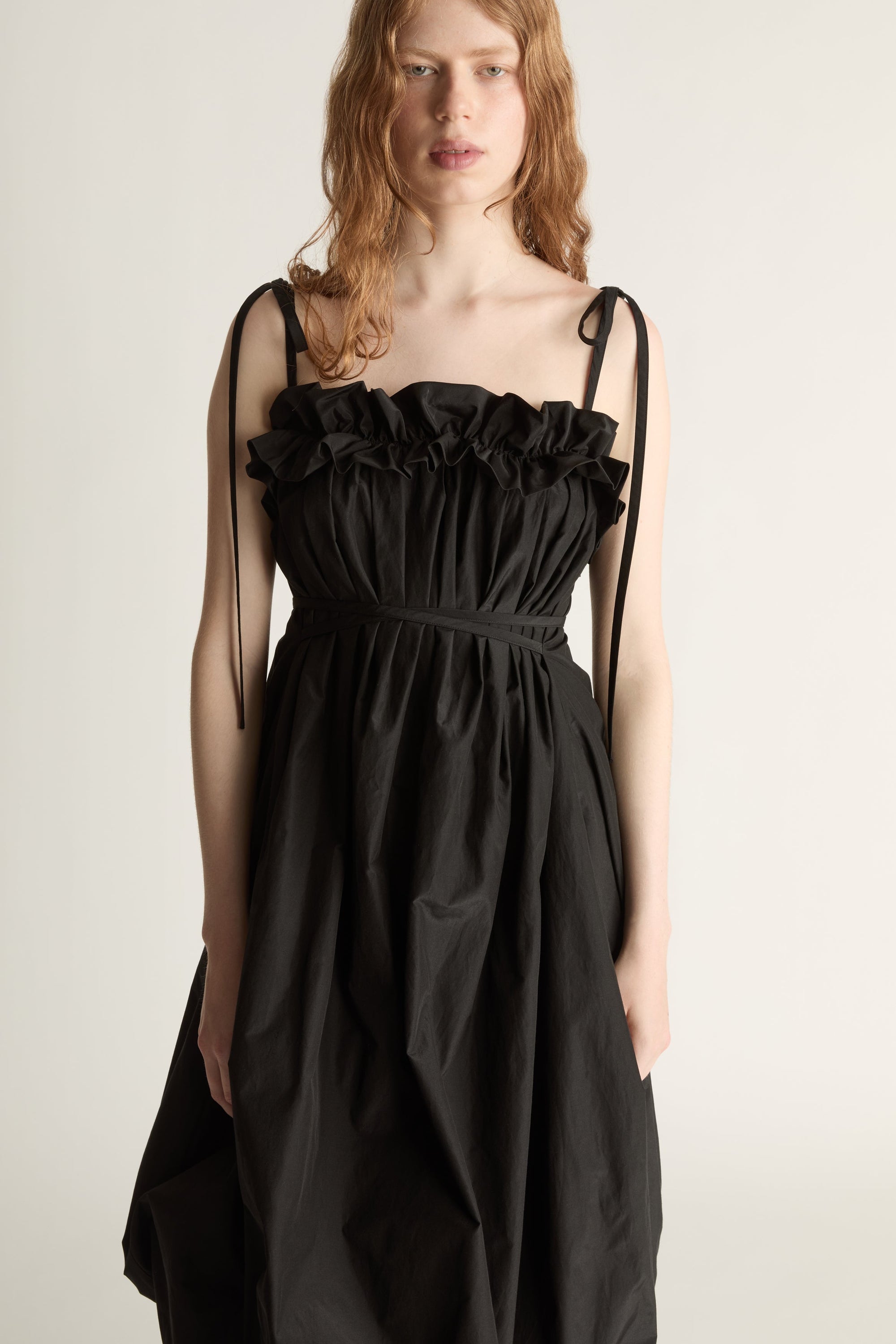 Close up of a woman wearing a black bubble style dress with a ruffle and tie shoulder straps against a white background.
