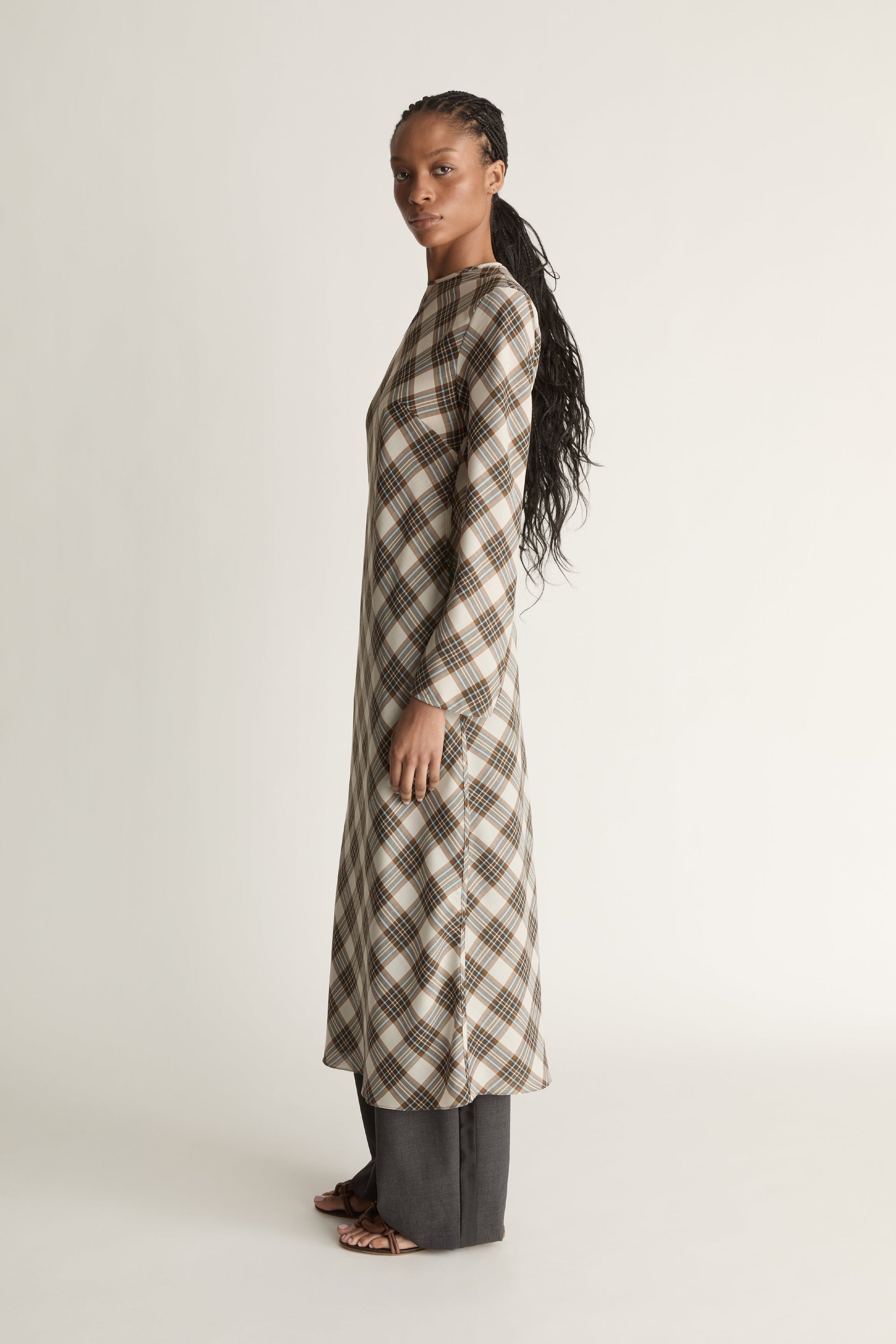 Side view of a woman wearing a long- sleeve midi length plaid dress on a white background