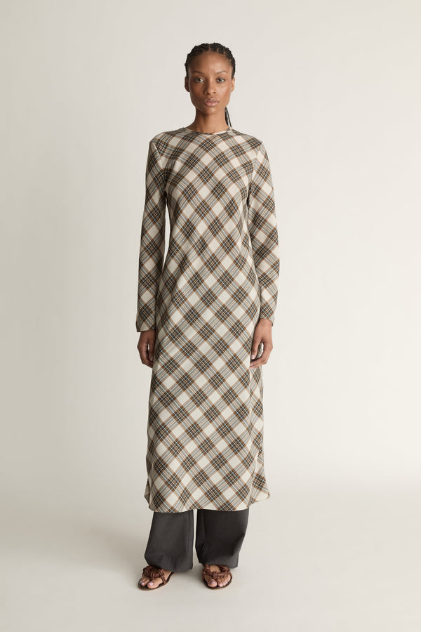 Front view of a woman wearing a long sleeve plaid midi length dress with grey trousers underneath and brown shoes on a white studio background.