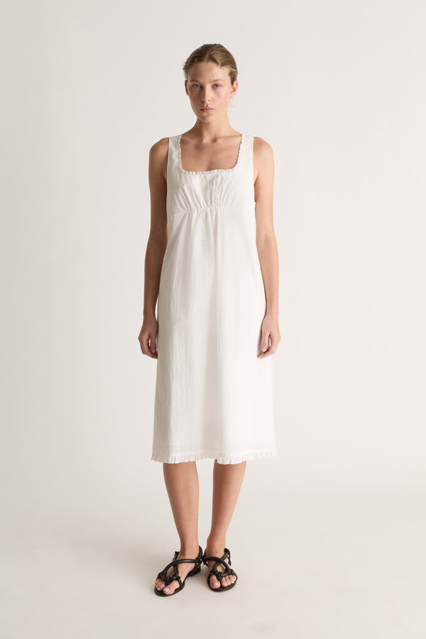Front view of a woman with hair pulled back in a white slip-style dress with frayed hem and black strappy sandals