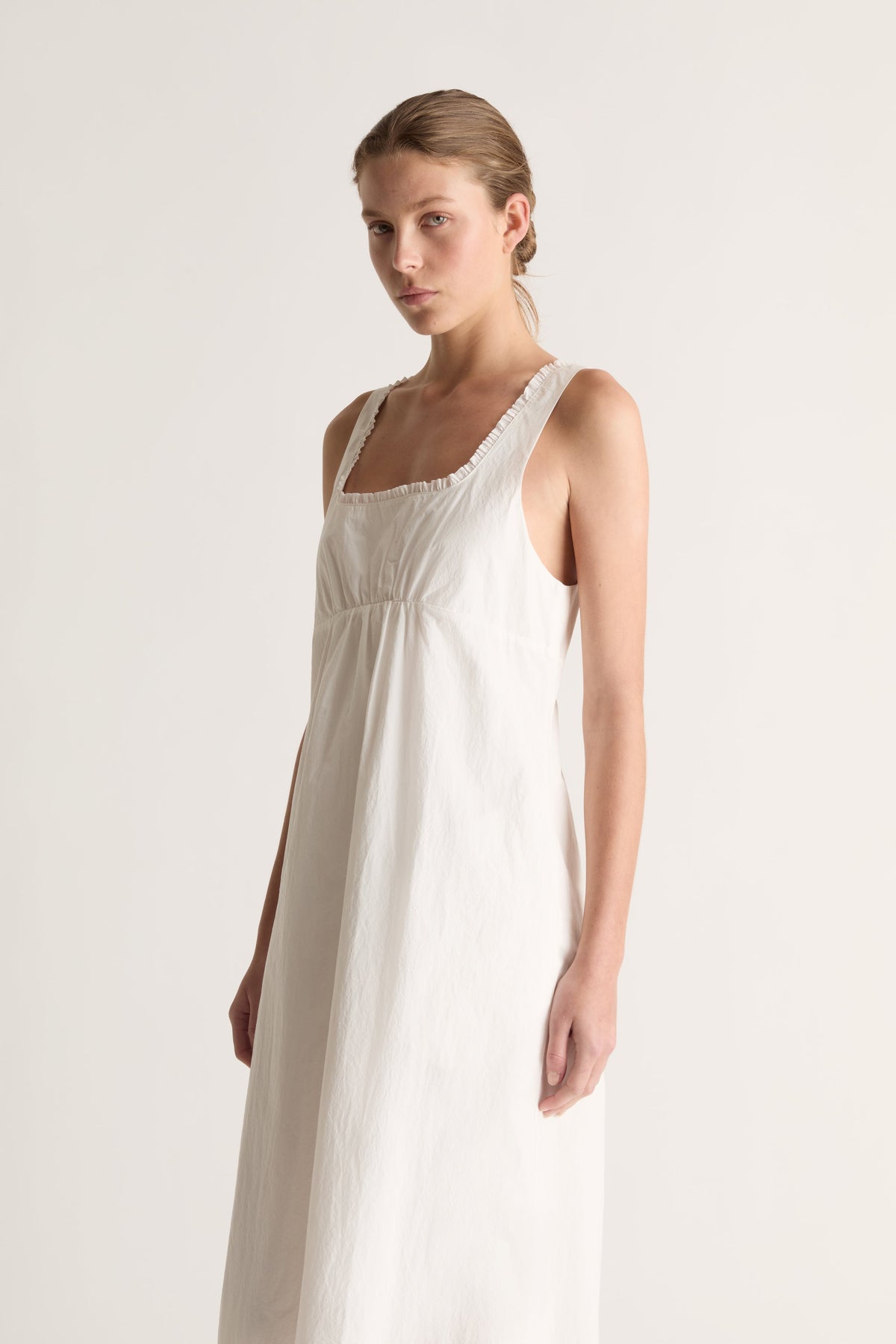 Angled close-up of a woman in a white empire-waist dress with ruffle-edged neckline and wide straps