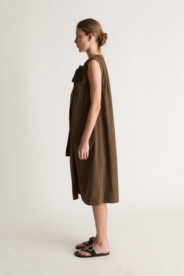 Side profile of a woman in a sleeveless khaki dress with black bow at the shoulder and black sandals