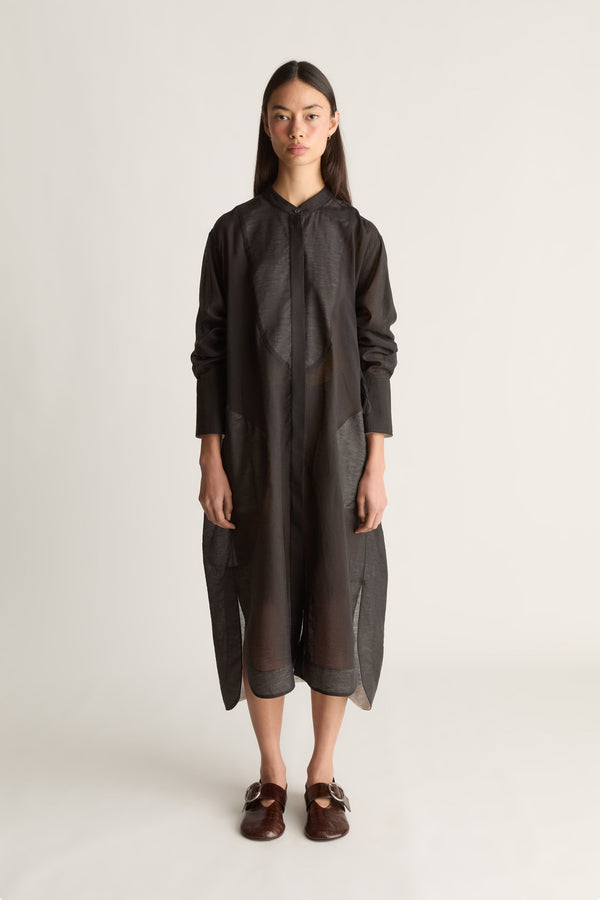 A model stands against a light gray studio background wearing a loose black long-sleeve dress with a high collar and draped front, paired with dark brown loafers.