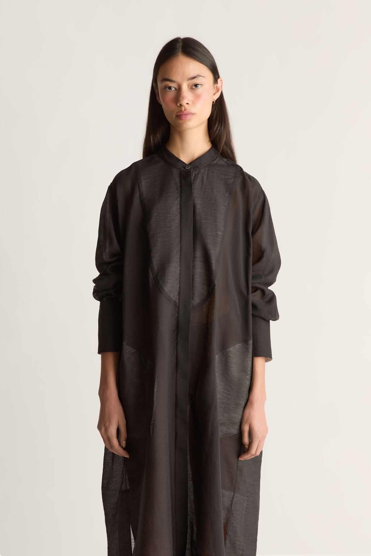 A model wearing a long, loose-fitting black sheer shirt dress with textured panel details, photographed against a plain light gray studio background.