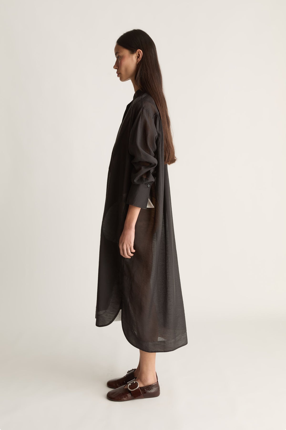 A woman stands in profile wearing a long, loose-fitting black dress with a high collar and long sleeves, paired with dark brown loafers, against a plain light background.