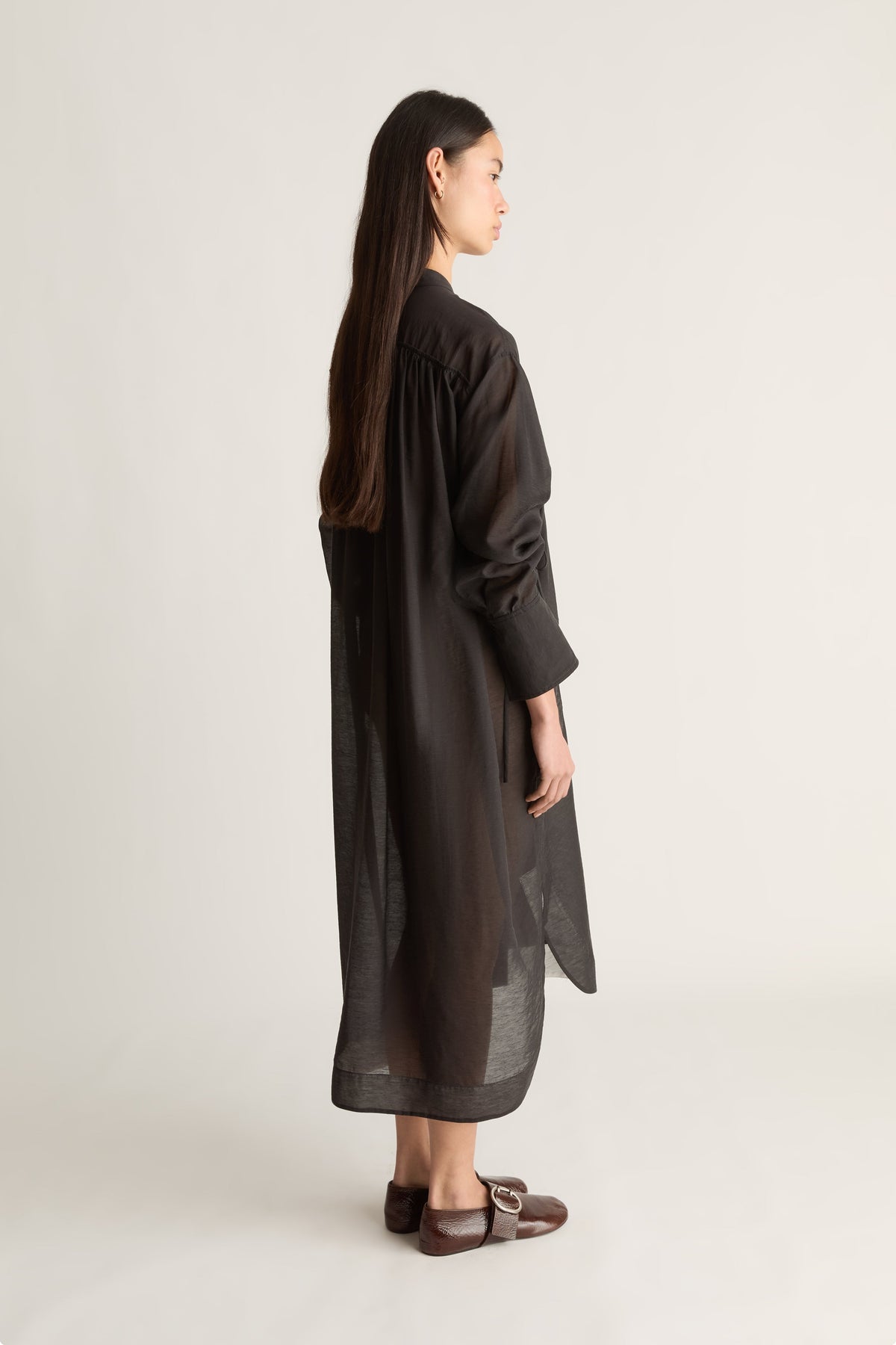 A woman shown from the back wearing a long, loose black dress or tunic with wide sleeves and a sheer textured fabric, paired with brown flat shoes, against a plain light gray studio background.