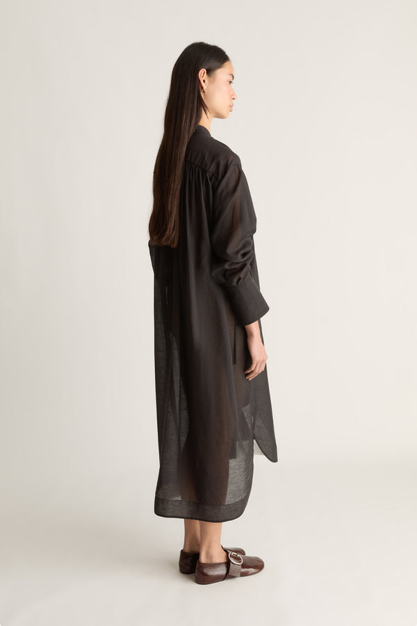 A woman shown from the back wearing a long, loose black dress or tunic with wide sleeves and a sheer textured fabric, paired with brown flat shoes, against a plain light gray studio background.