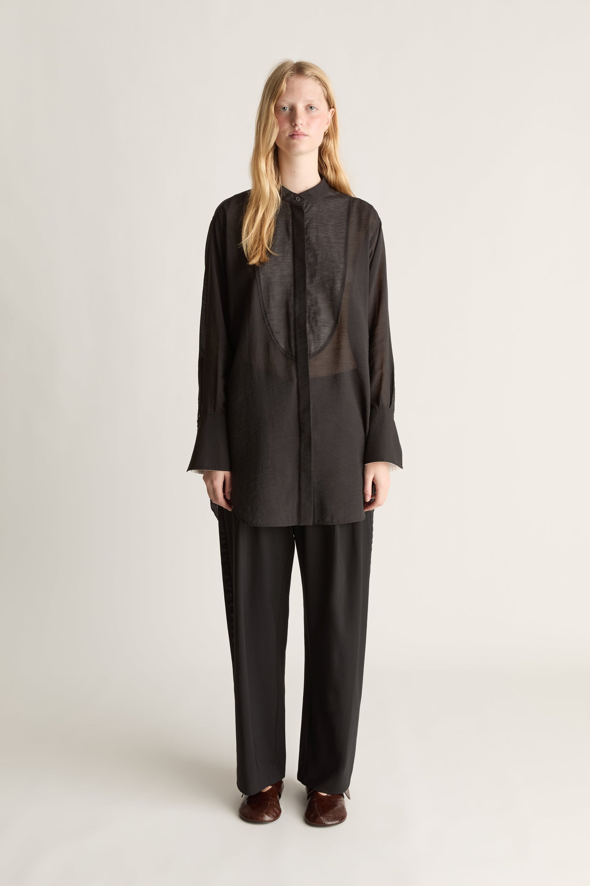 A model stands against a light gray studio background wearing a black long-sleeve button-front top with sheer front panels and matching black wide-leg pants.