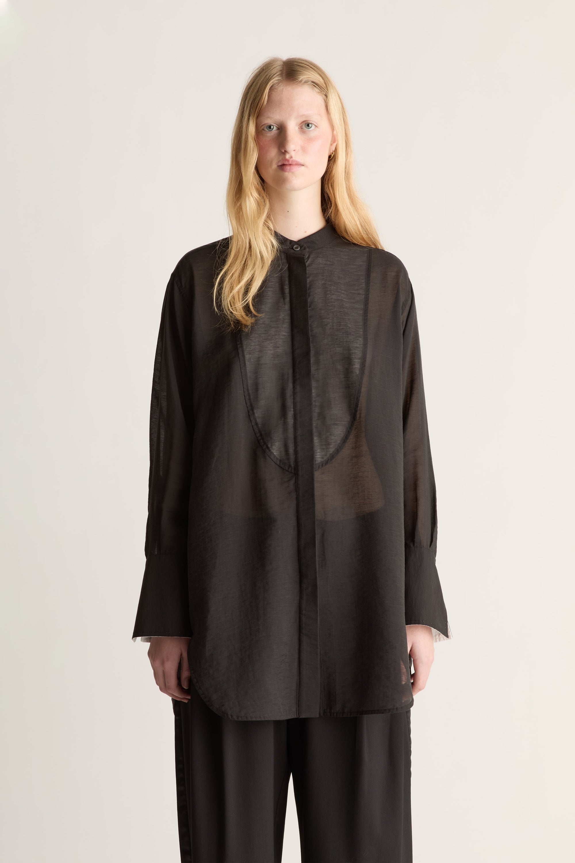 A close up of a model wearing  a loose, semi-sheer black long-sleeve blouse with a high collar and matching black trousers, standing against a plain light beige studio background.