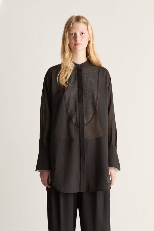 A close up of a model wearing  a loose, semi-sheer black long-sleeve blouse with a high collar and matching black trousers, standing against a plain light beige studio background.