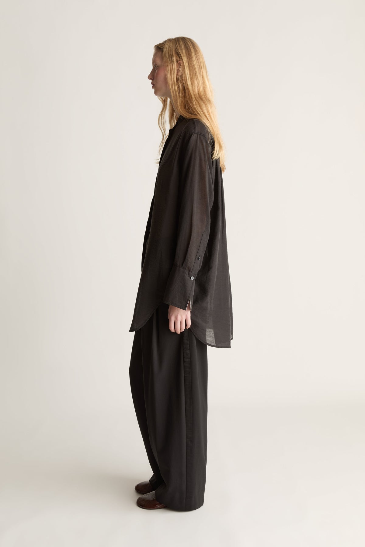 A woman with long blonde hair stands in profile wearing a loose black long-sleeve top and matching wide-leg pants against a plain light gray studio background.