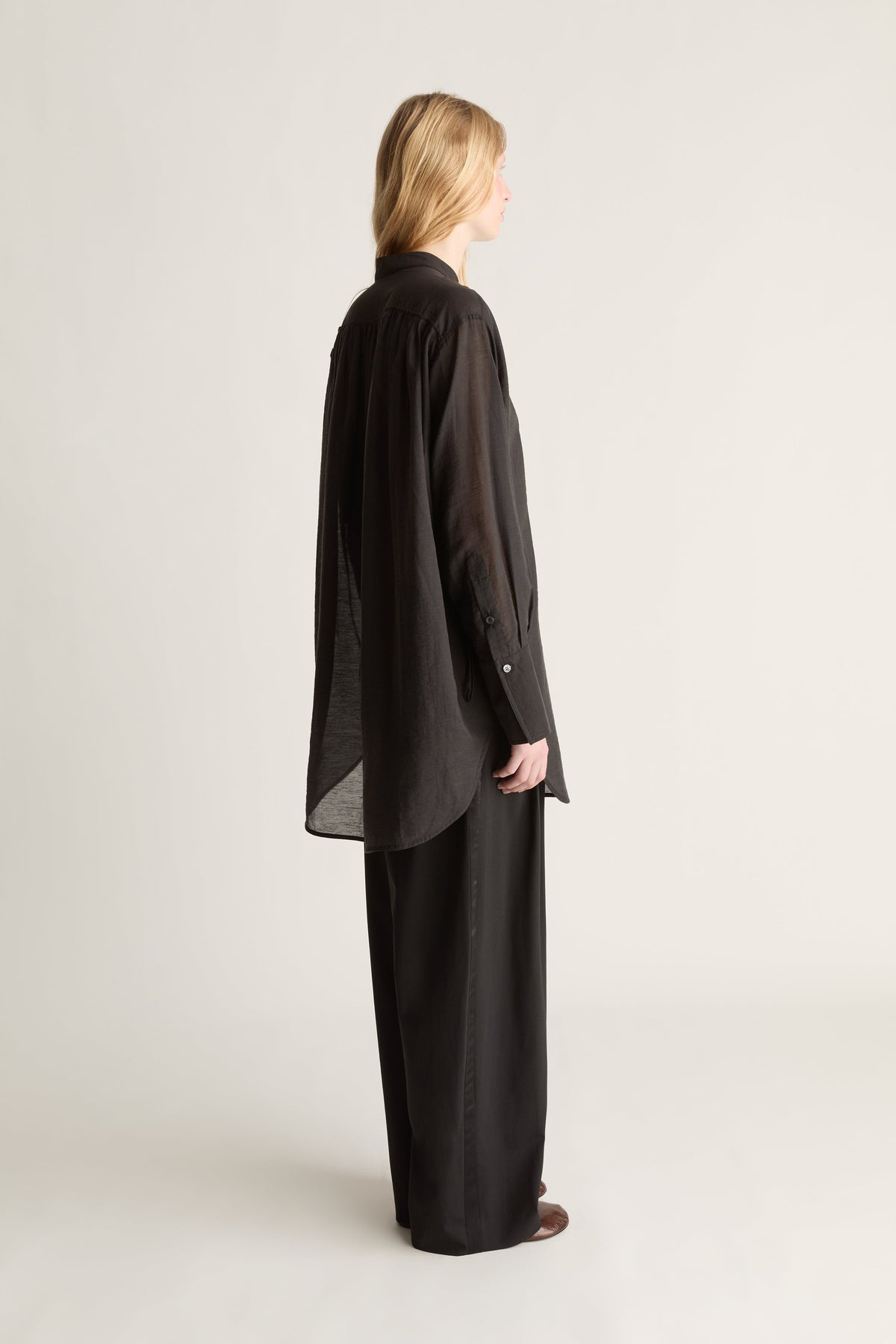 Back profile image of a woman wearing a loose black long-sleeve top and matching wide-leg black pants against a plain light gray studio background.