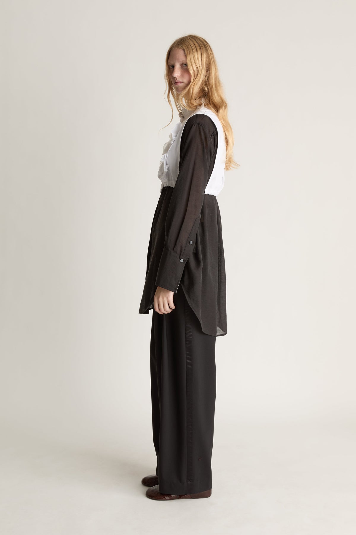 Side view of a woman wearing a black shirt and pants with a white frilled vest against a white studio background.