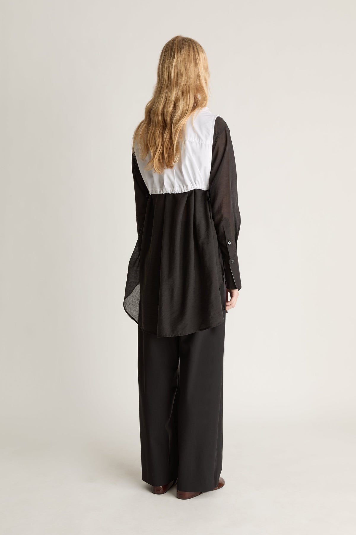 Back view of a person wearing a black long sleeve shirt and black trousers with a white frilled dickey vest, against a white studio background.