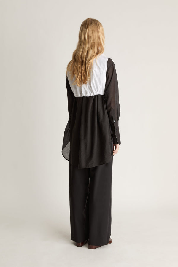Back view of a person wearing a black long sleeve shirt and black trousers with a white frilled dickey vest, against a white studio background.