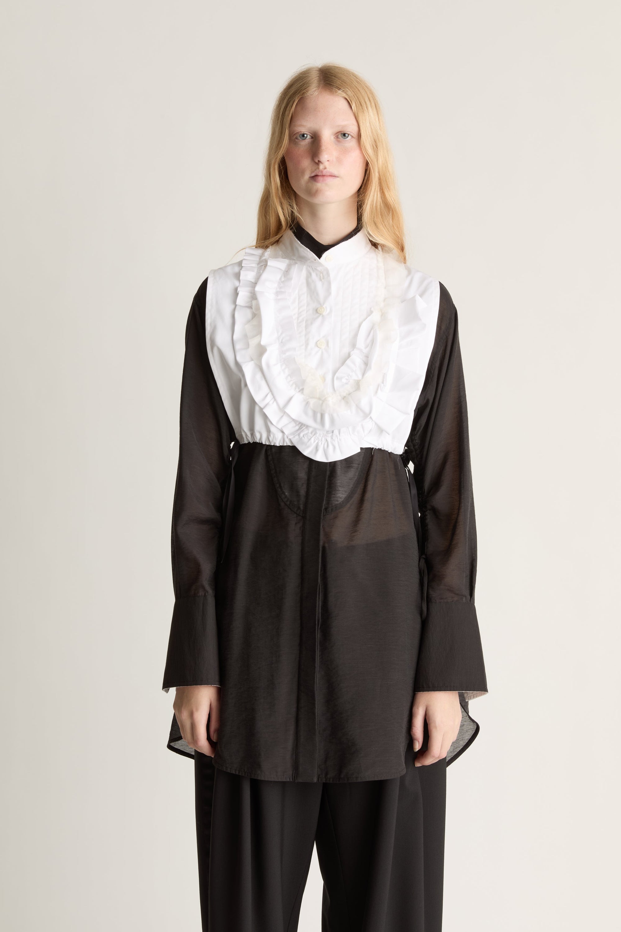 Front view of a person wearing a black long sleeve shirt and black trousers with a white frilled dickey vest, against a white studio background.