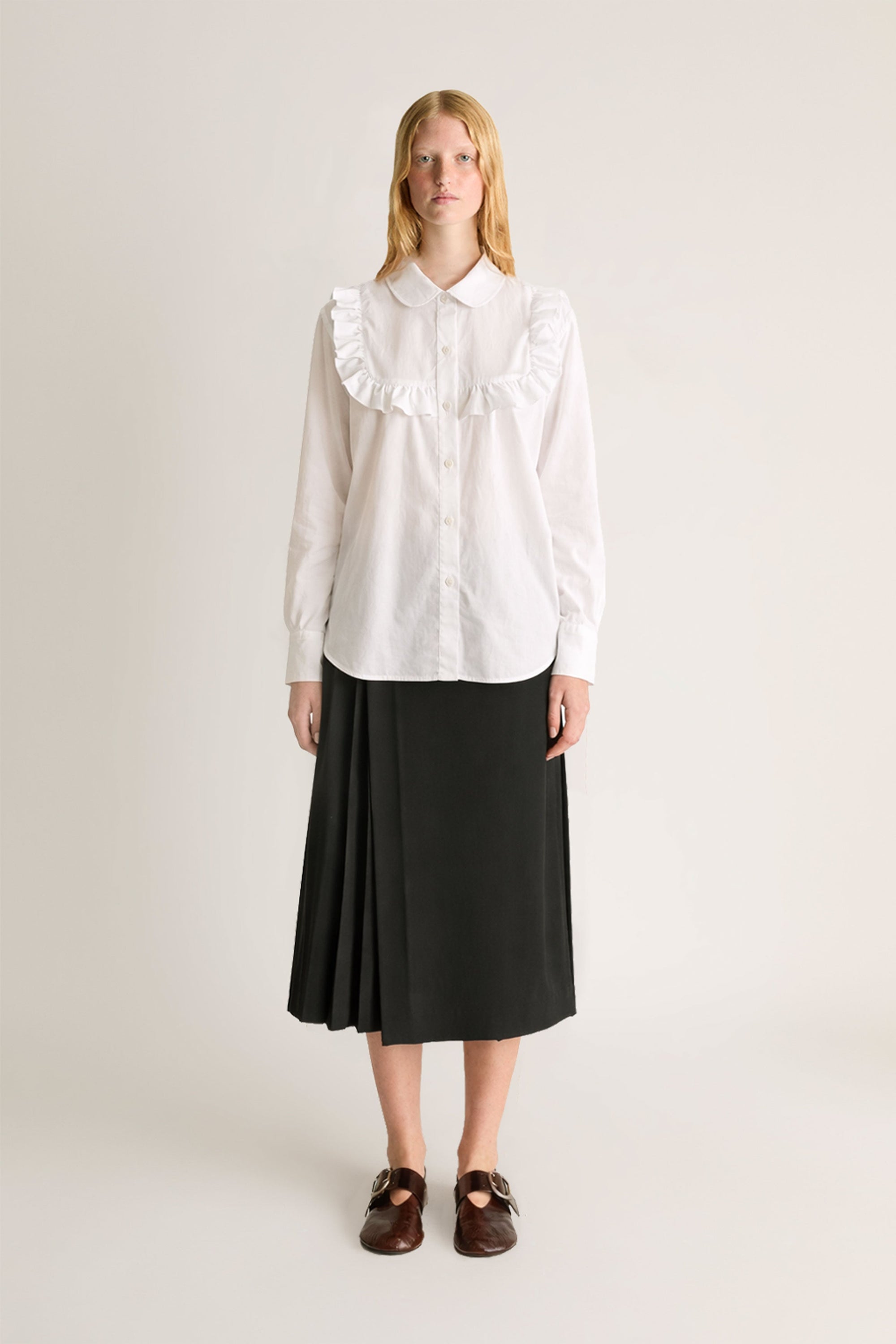 Person wearing a white blouse with ruffled collar and a black skirt on a plain background