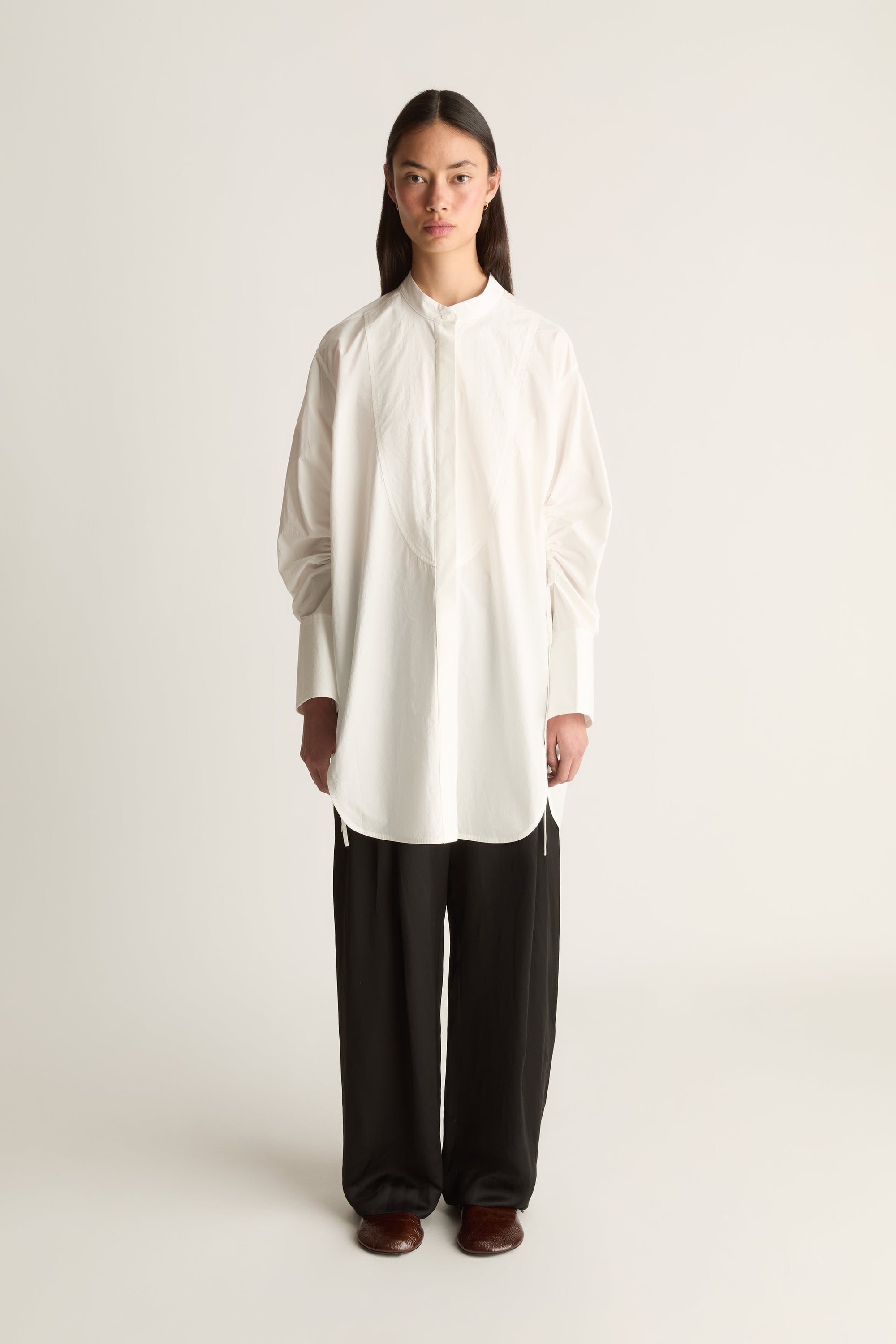 A model stands against a plain light background wearing a loose white long-sleeve button-up shirt with a high collar and black wide-leg pants.