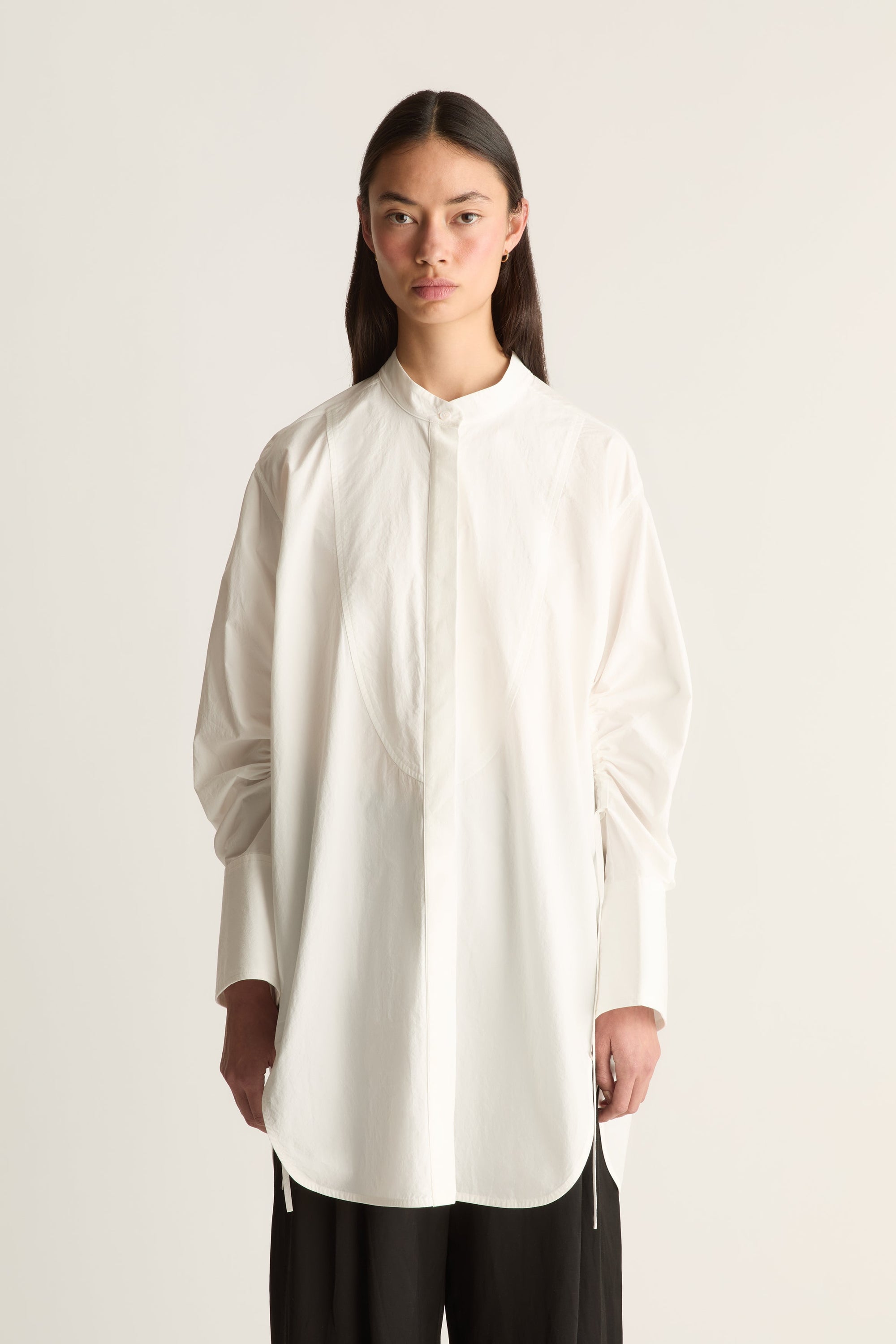 Close up of a model wearing an oversized white long-sleeve blouse with a high collar and gathered sleeves, paired with black pants, against a plain off-white studio background.