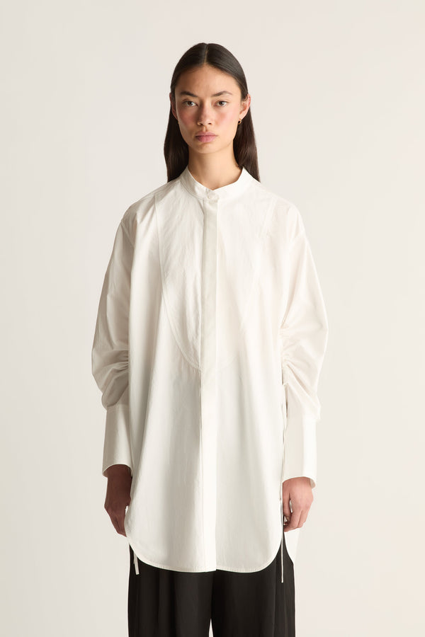Close up of a model wearing an oversized white long-sleeve blouse with a high collar and gathered sleeves, paired with black pants, against a plain off-white studio background.