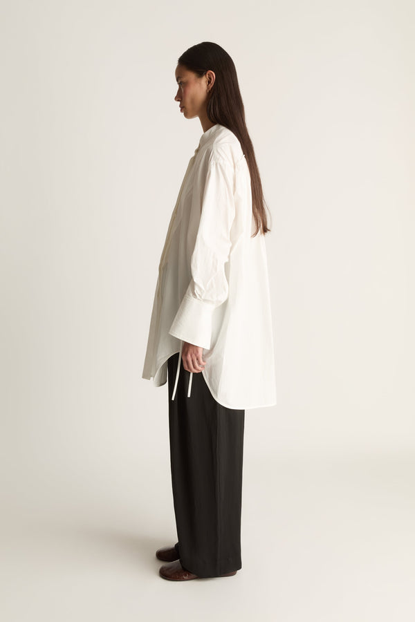 A woman stands in profile wearing an oversized white long-sleeve blouse with a high-low hem and black wide-leg trousers against a plain light background.