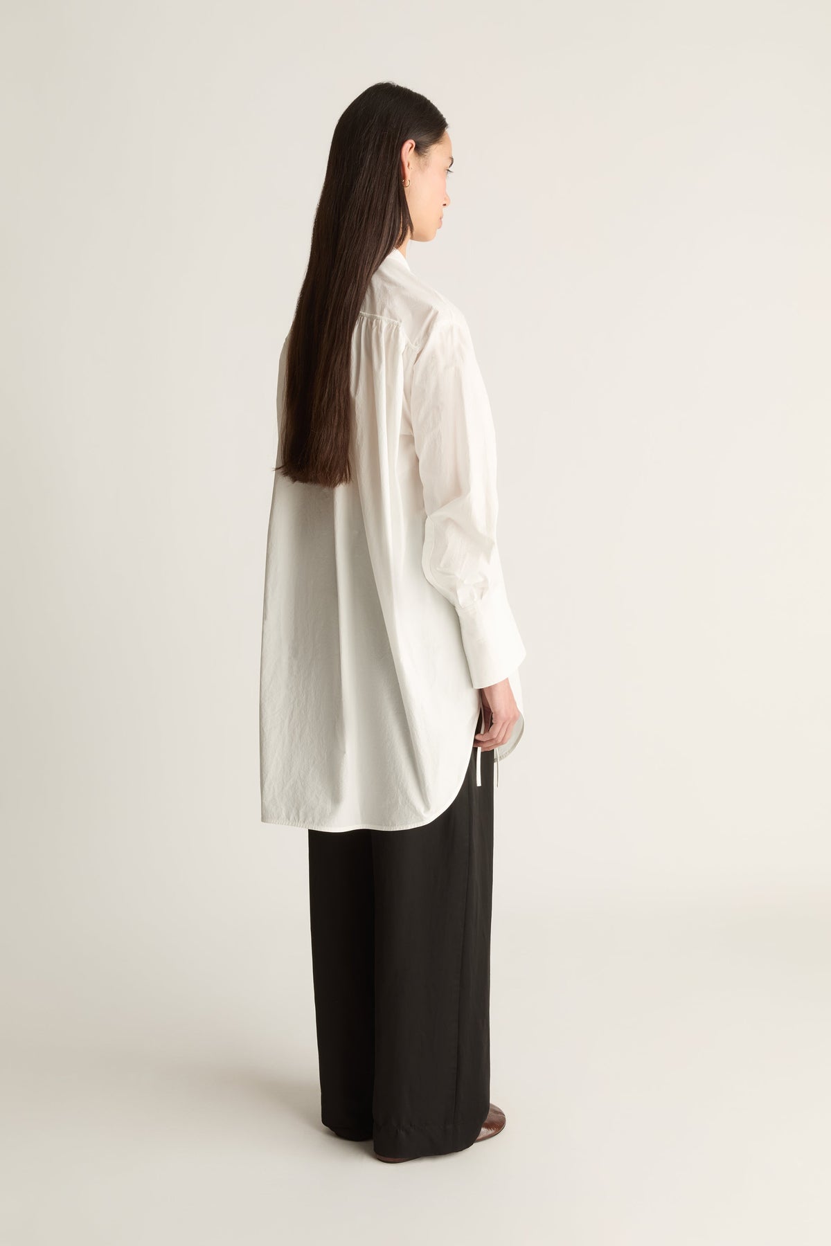 A woman shown from the back wearing a loose white long-sleeve blouse and black wide-leg pants against a plain light gray studio background.