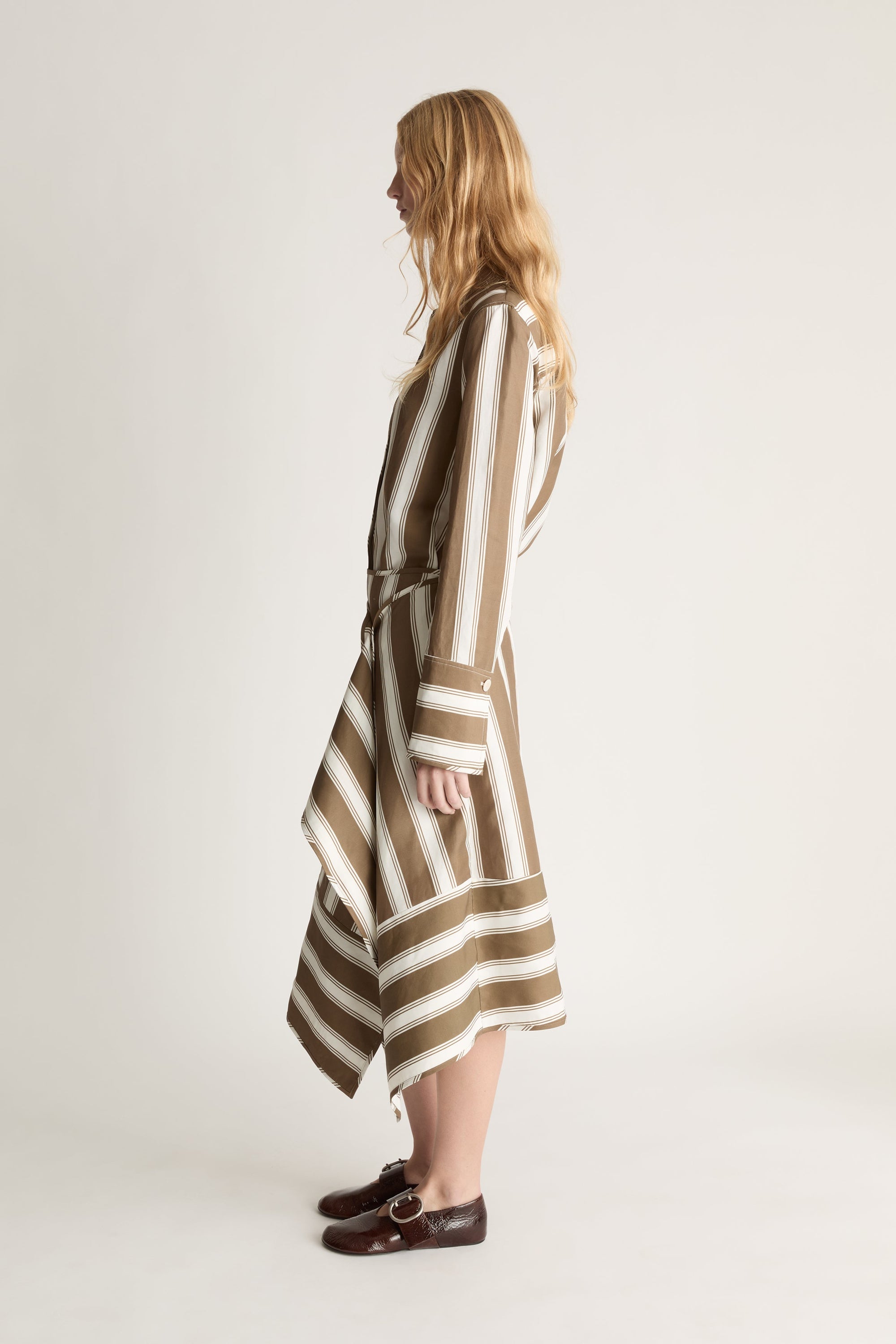 Profile view of a woman wearing a brown and white striped button up shirt and a matching wrap skirt against a white studio background. 