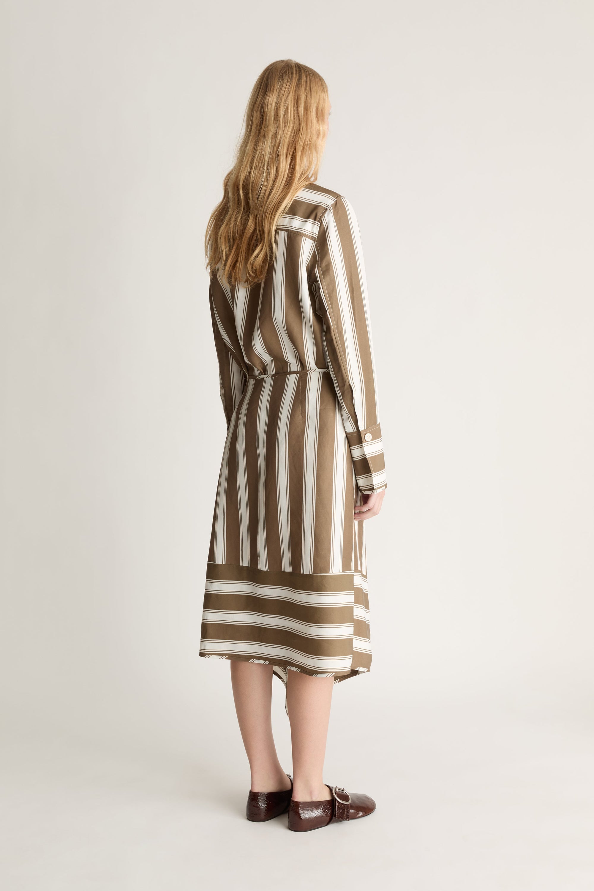 Back view of a woman wearing a brown and white striped button up shirt and skirt against a white background. 