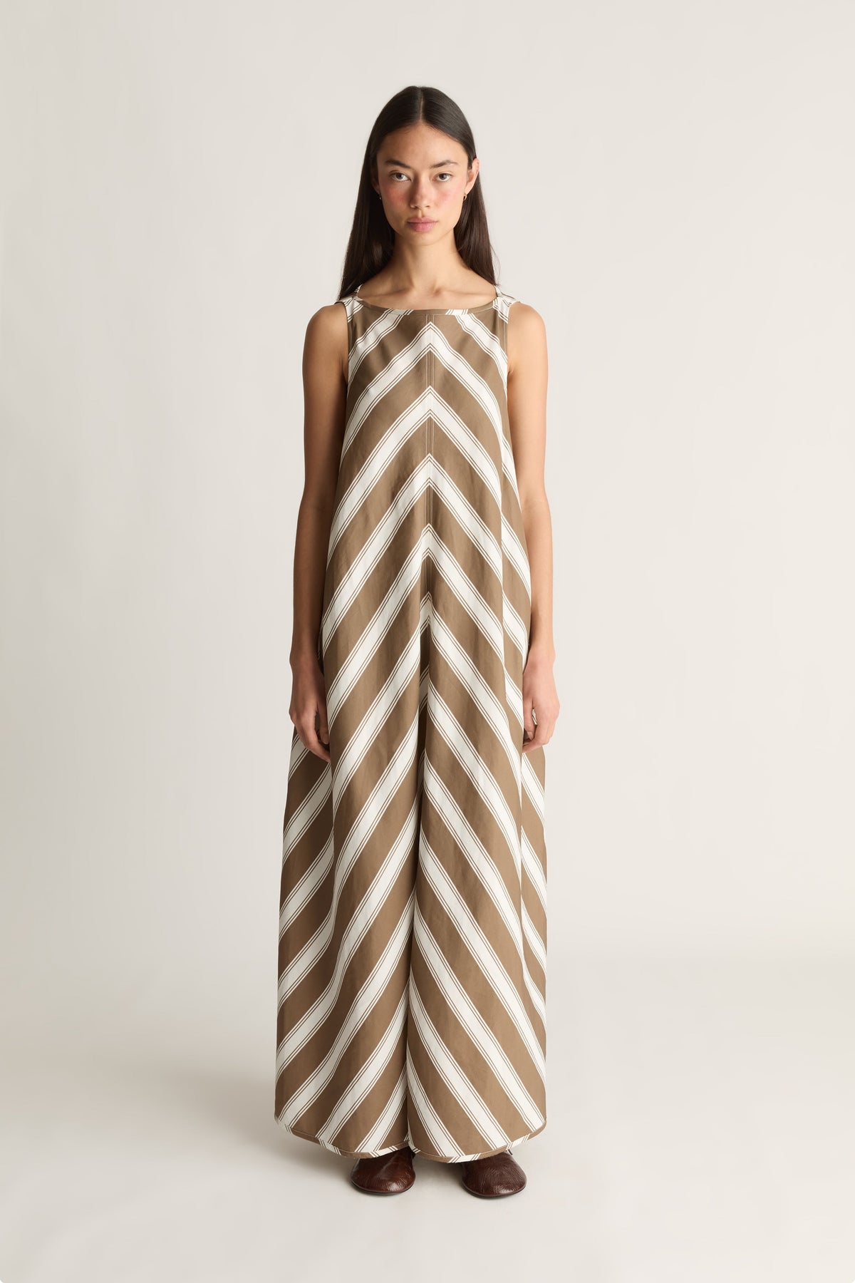 A woman stands against a plain light gray studio background wearing an A-linefull length dress with bold diagonal stripes in warm tan and off-white, short against a white studio background. 