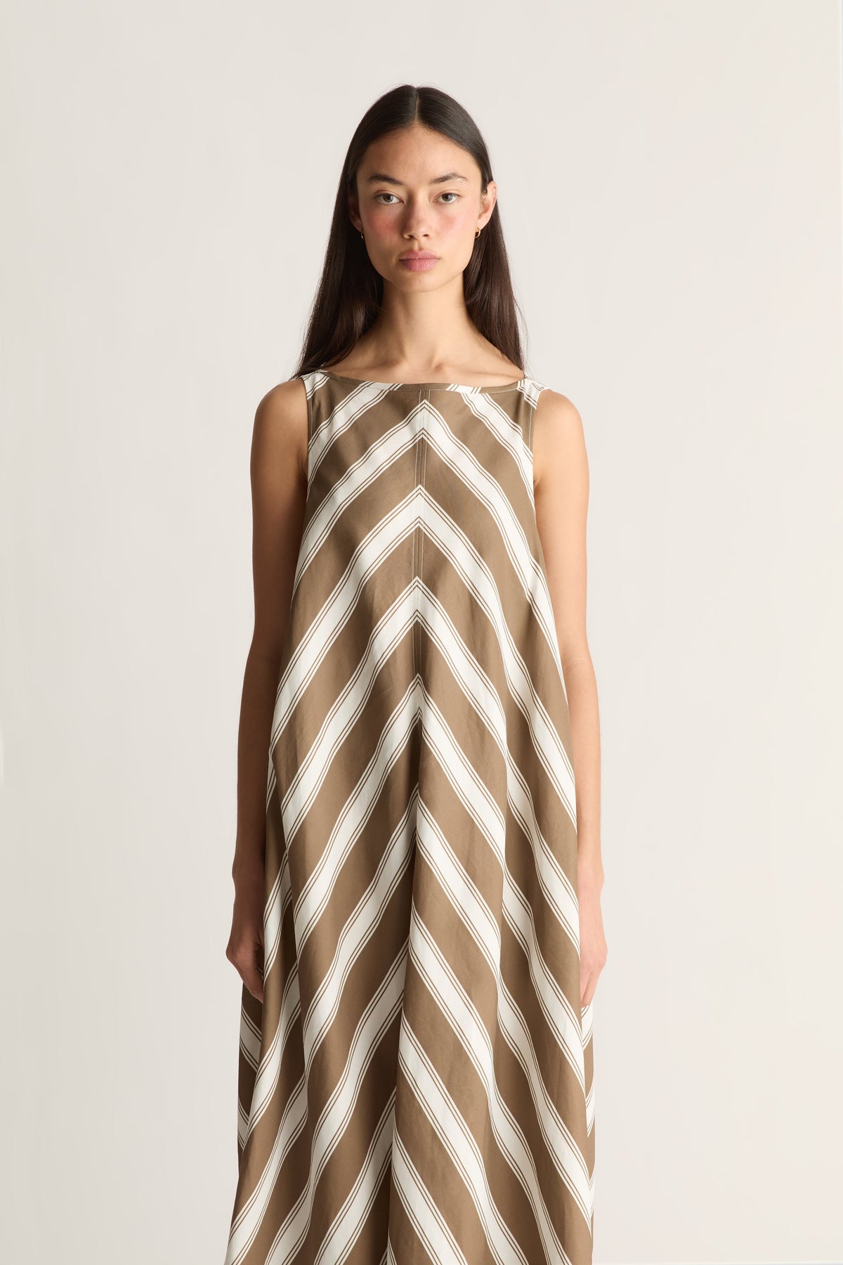 A woman modeling a sleeveless maxi dress with a brown and off-white chevron pattern against a plain light background.