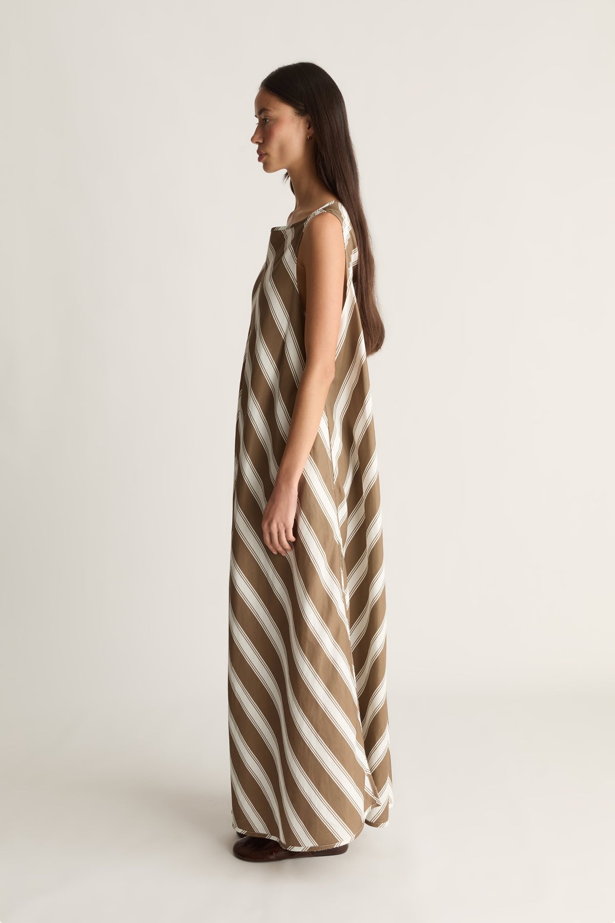 A woman modeling a sleeveless maxi dress with diagonal brown and off-white stripes, shown in profile against a plain light background.