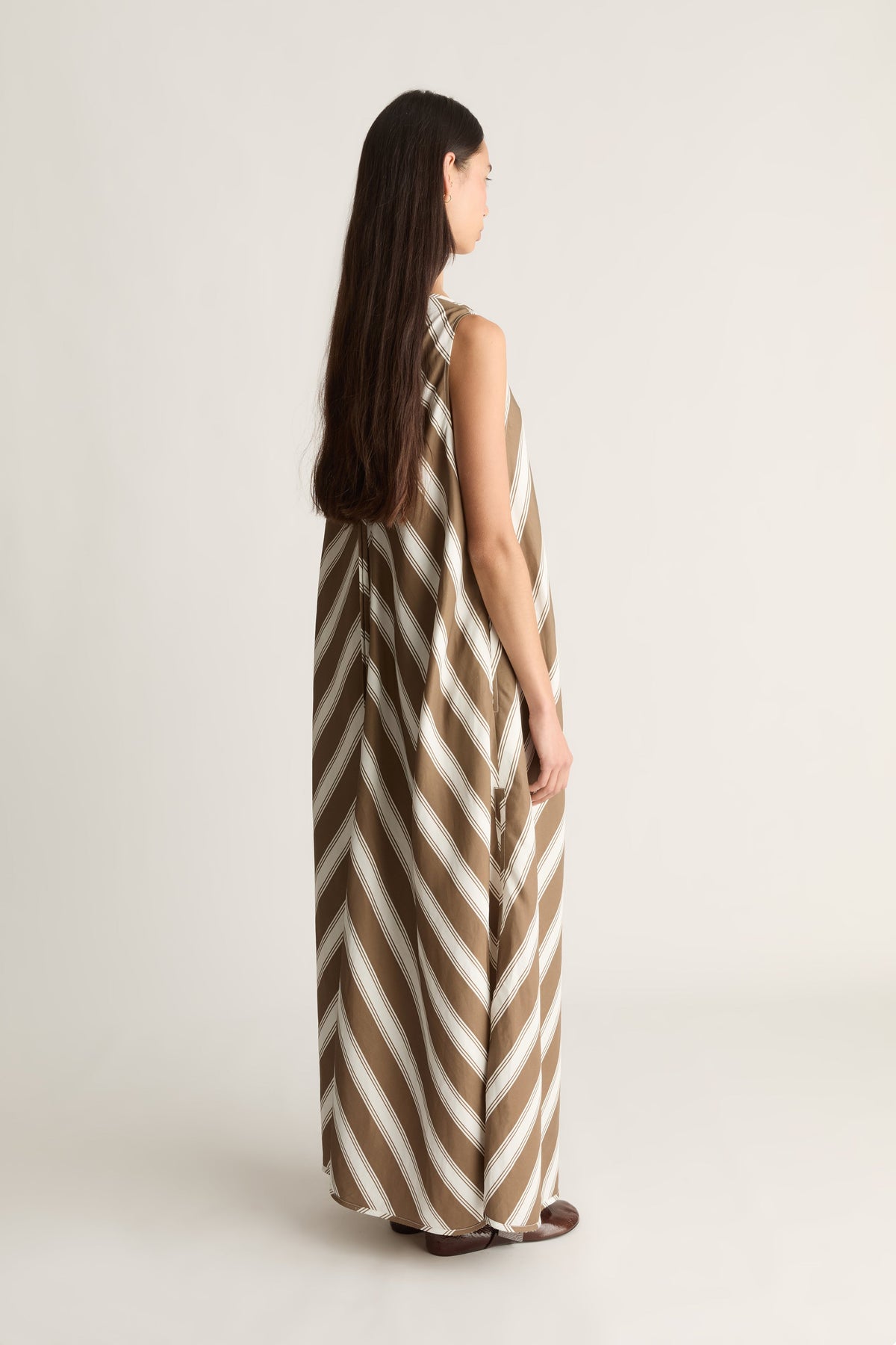 A woman stands in profile wearing a sleeveless, floor-length dress with diagonal taupe-brown and off-white stripes against a plain light beige studio background.