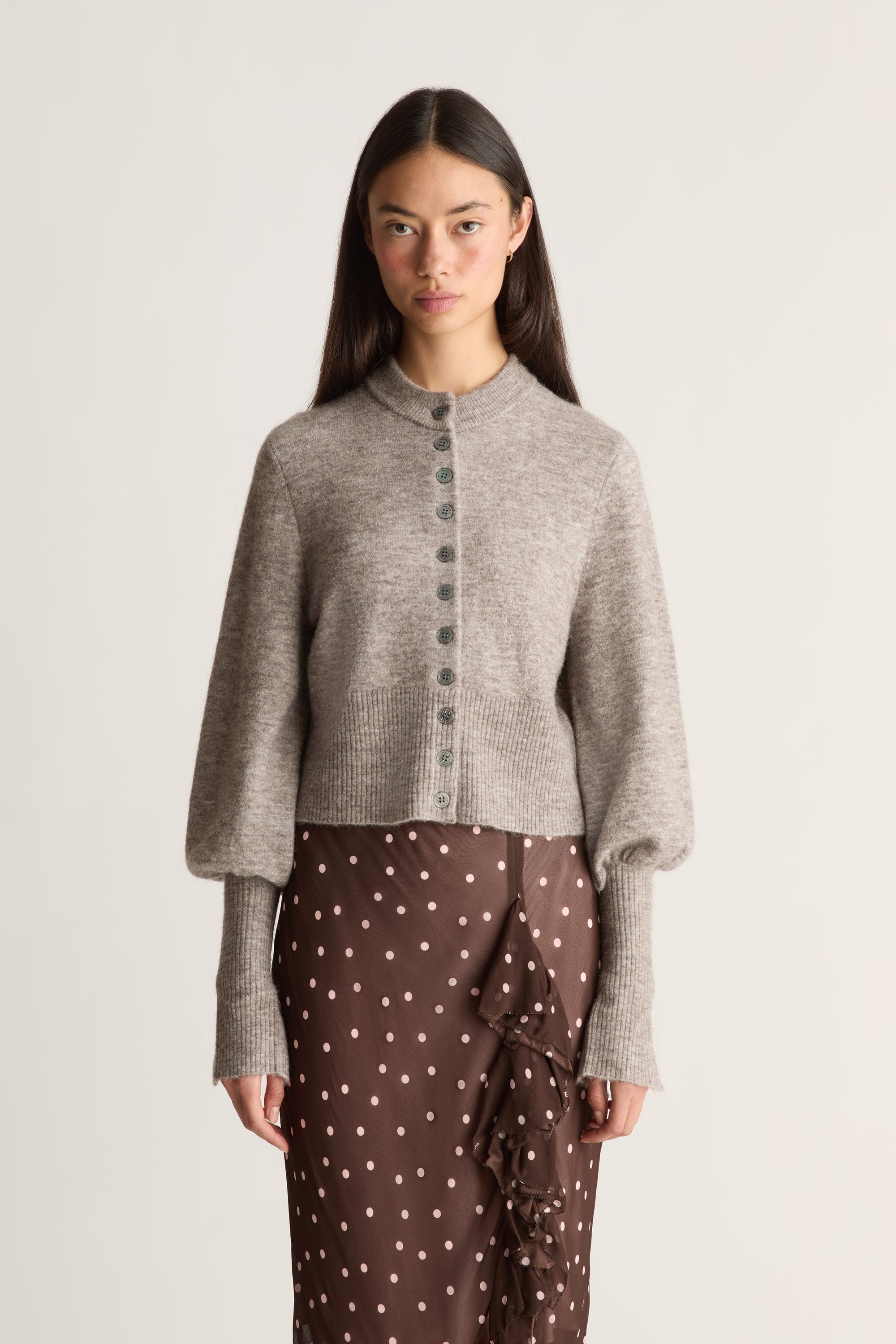 Close up of a woman wearing a gray knit cardigan over a brown skirt with white polka dots on a light background