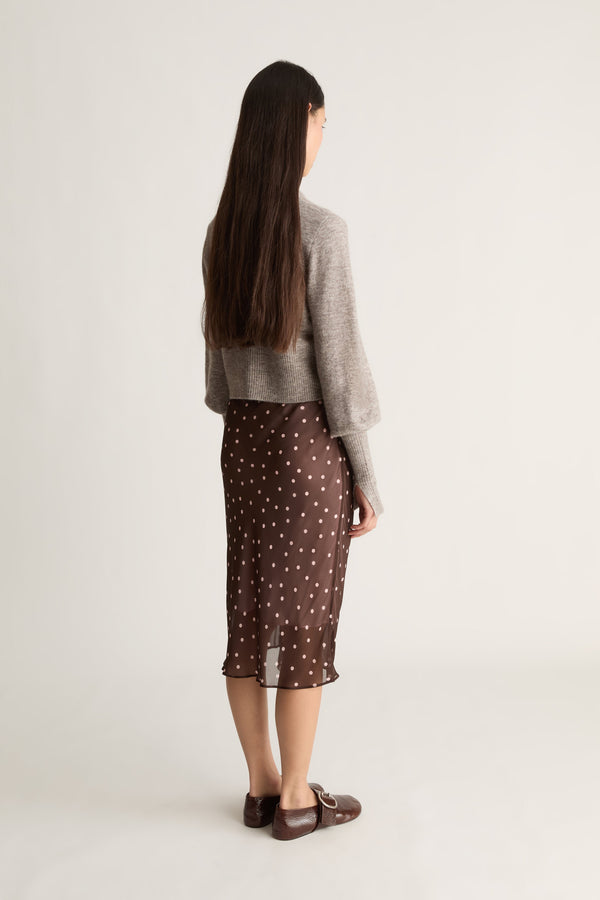 Back view of a woman wearing a gray cardigan and brown skirt with white polka dots on a white background